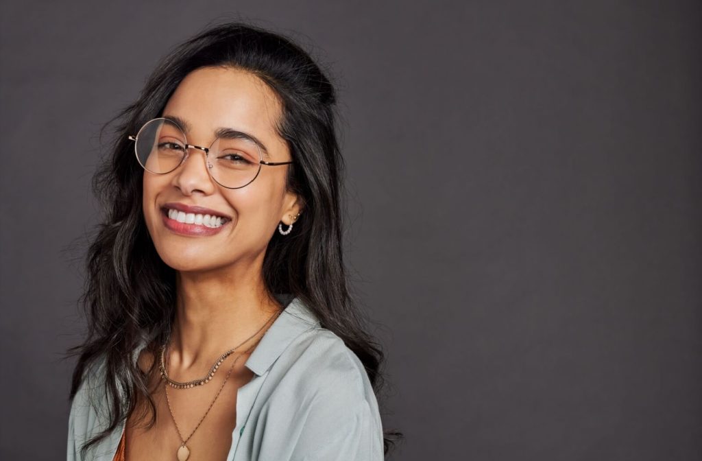 Smiling woman wearing round Asian fit eyeglasses with a thin metal frame, standing against a dark background.