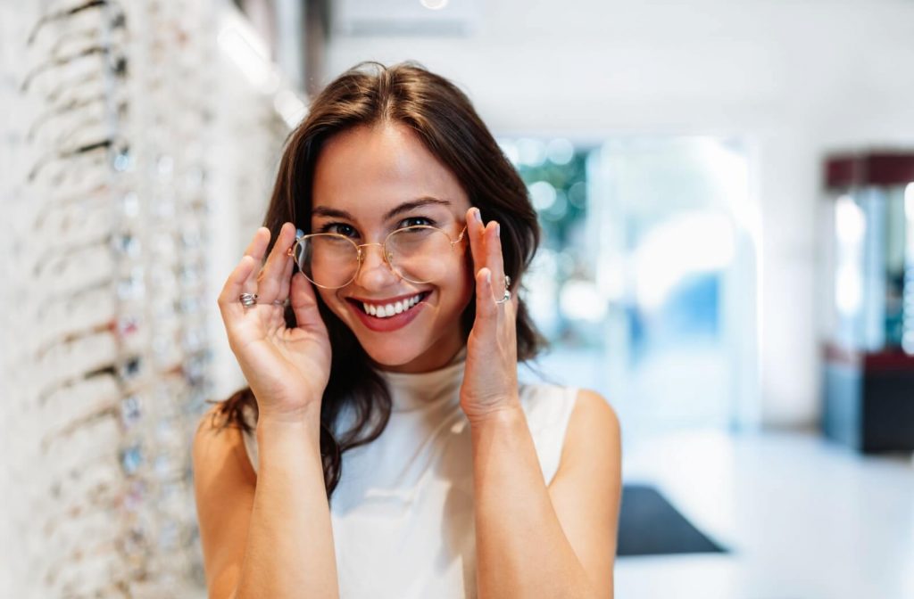 A smiling woman in an optical boutique adjusting a pair of gold-rimmed eyeglasses on her face while looking at her reflection.