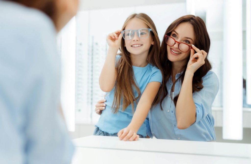 A girl and her mother smiling in front of a mirror while trying on eyeglasses at an optical store.