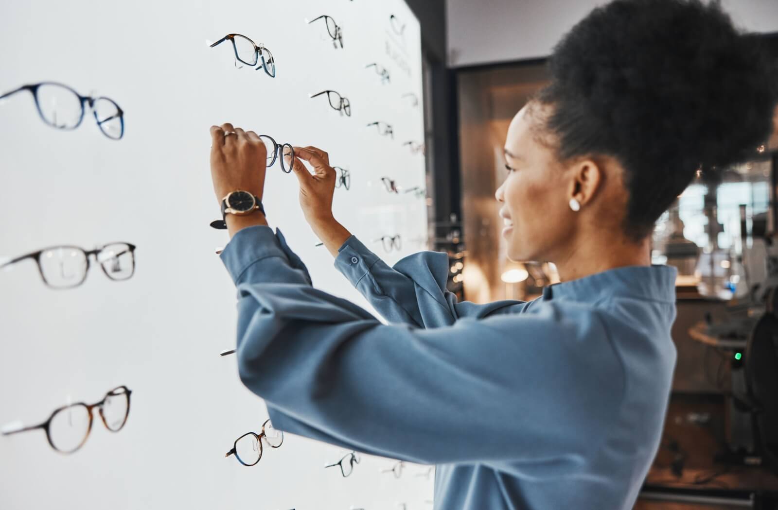 A person browsing a display wall of eyeglass frames, holding up a pair to examine them in a brightly lit optical store.