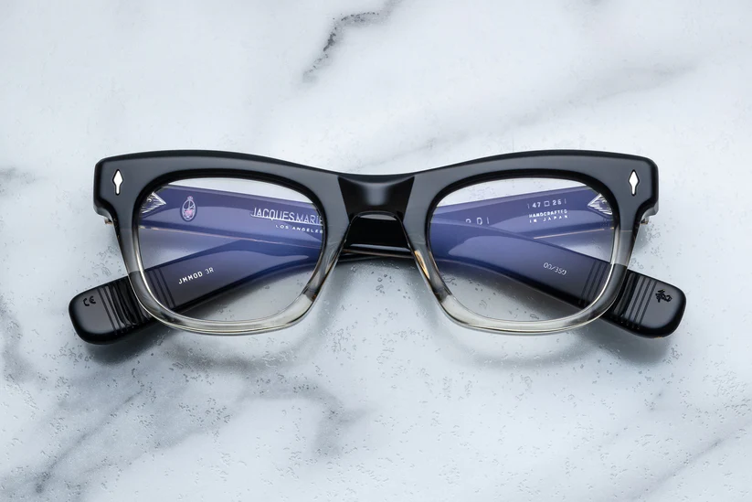 A pair of bold, square Jacques Marie Mage eyeglasses with a black-to-clear gradient acetate frame, displayed face-up on a smooth white marble surface.