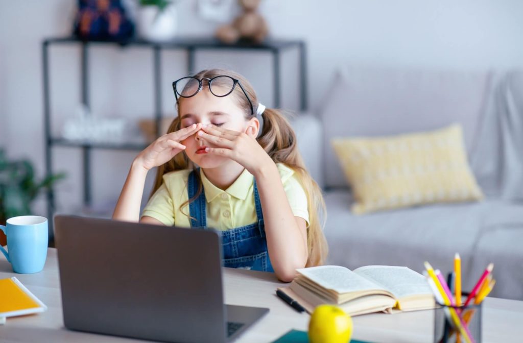 Child rubbing tired eyes while using a laptop for schoolwork at home, illustrating digital eye strain in children and the importance of regular pediatric eye exams.