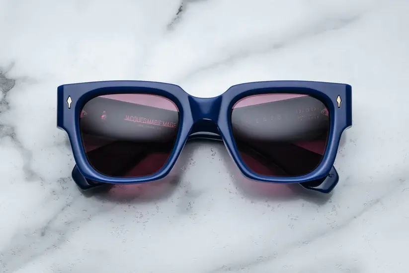Luxury Jacques Marie Mage ENZO sunglasses with thick navy blue frames and dark lenses, showcased on a marble surface as part of a style guide for first-time buyers.
