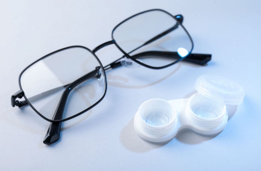 A pair of black-framed eyeglasses resting next to an open white contact lens case on a light surface.
