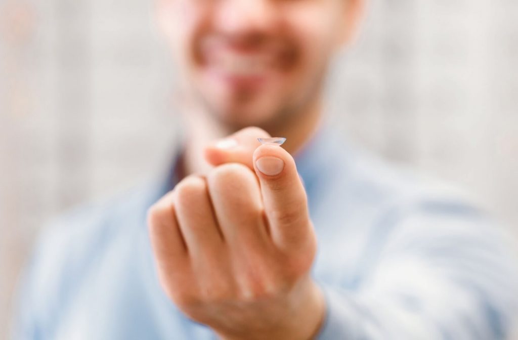 A close-up of a contact lens resting on an individual's fingertip with a blurred, smiling person in the background.
