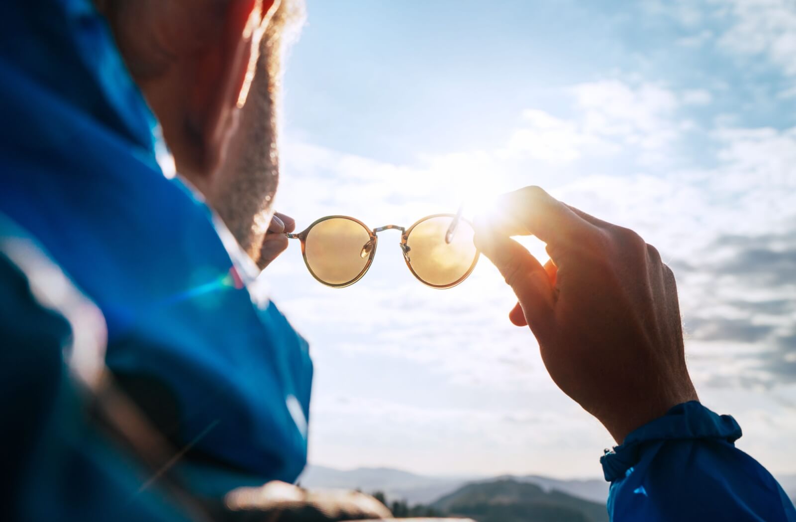 An over-the-shoulder view of a person holding up a pair of round, yellow-tinted sunglasses toward a bright, sunlit sky to block glare.