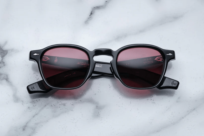 A top-down view of Jacques Marie Mage sunglasses featuring a thick black acetate frame and dark rose-tinted lenses, displayed on a white marble surface.