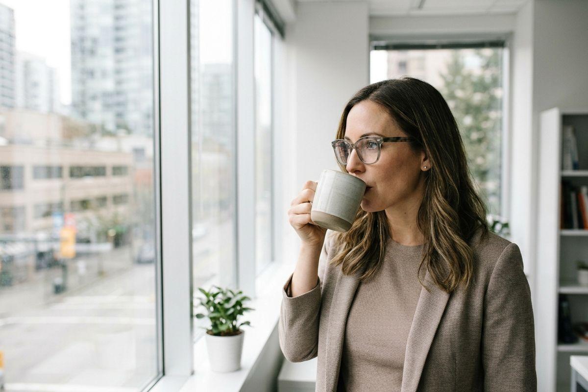 Person in glasses and beige blazer drinking from a mug, looking out an office window over city buildings.