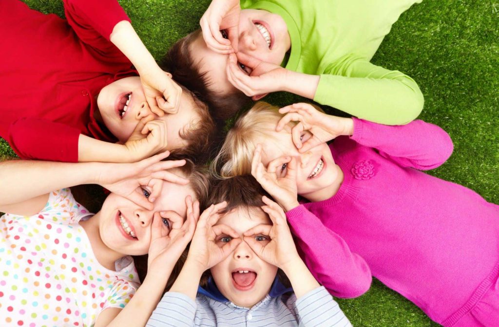 A group of children laying on the ground, making a glasses shape over their eyes using their hands.