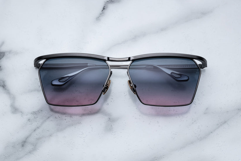 Geometric sunglasses with dark metal browline frames and gradient blue-to-purple tinted lenses resting on a marble surface.